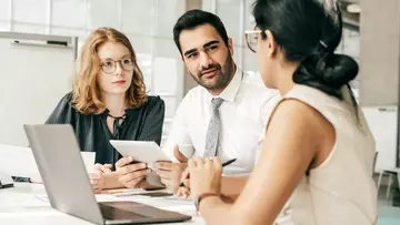 Three business people, a man in the middle of two women and talking at a table over laptops and tablets.