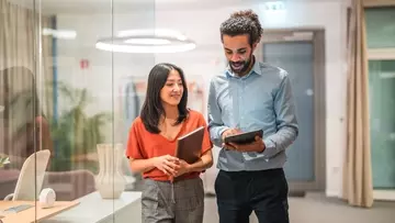 two employees walking down a hall and talking to each other while holding tablets.