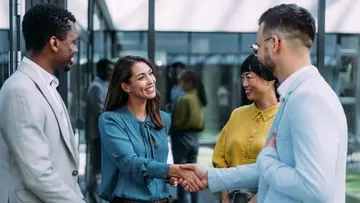 Four business people in front of a modern office building. A man and a woman are shaking hands.