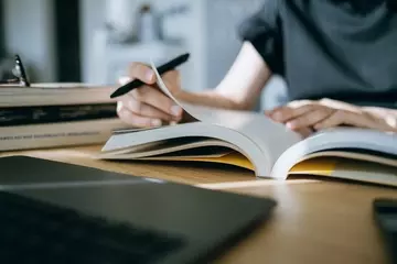 A woman's torso is shown from the neck down. She is sititng behind a table reading a book and annotating with a pen. 