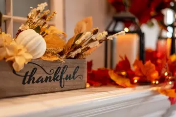 A mantle decorated for Thanksgiving with pumpkins, fall leaves, candles and a box that reads "thankful" in script.