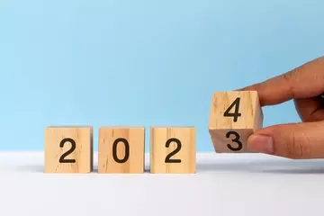 Blocks that read 2 0 2 3 are displayed on a blue backdrop. A hand reaches in from right to flip the 3 block to 4 so it reads 2024.