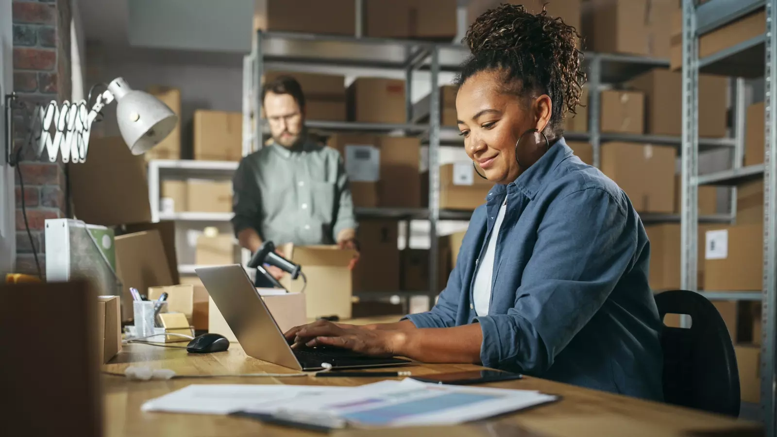 Diverse Male and Female Warehouse Inventory Managers Talking, Using Laptop Computer and Checking Retail Stock. Rows of Shelves Full of Cardboard Box Packages in the Background. 