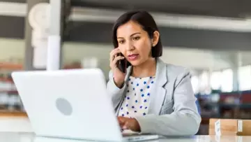 Female business woman in open plan office of a small business in front of a laptop and on the phone